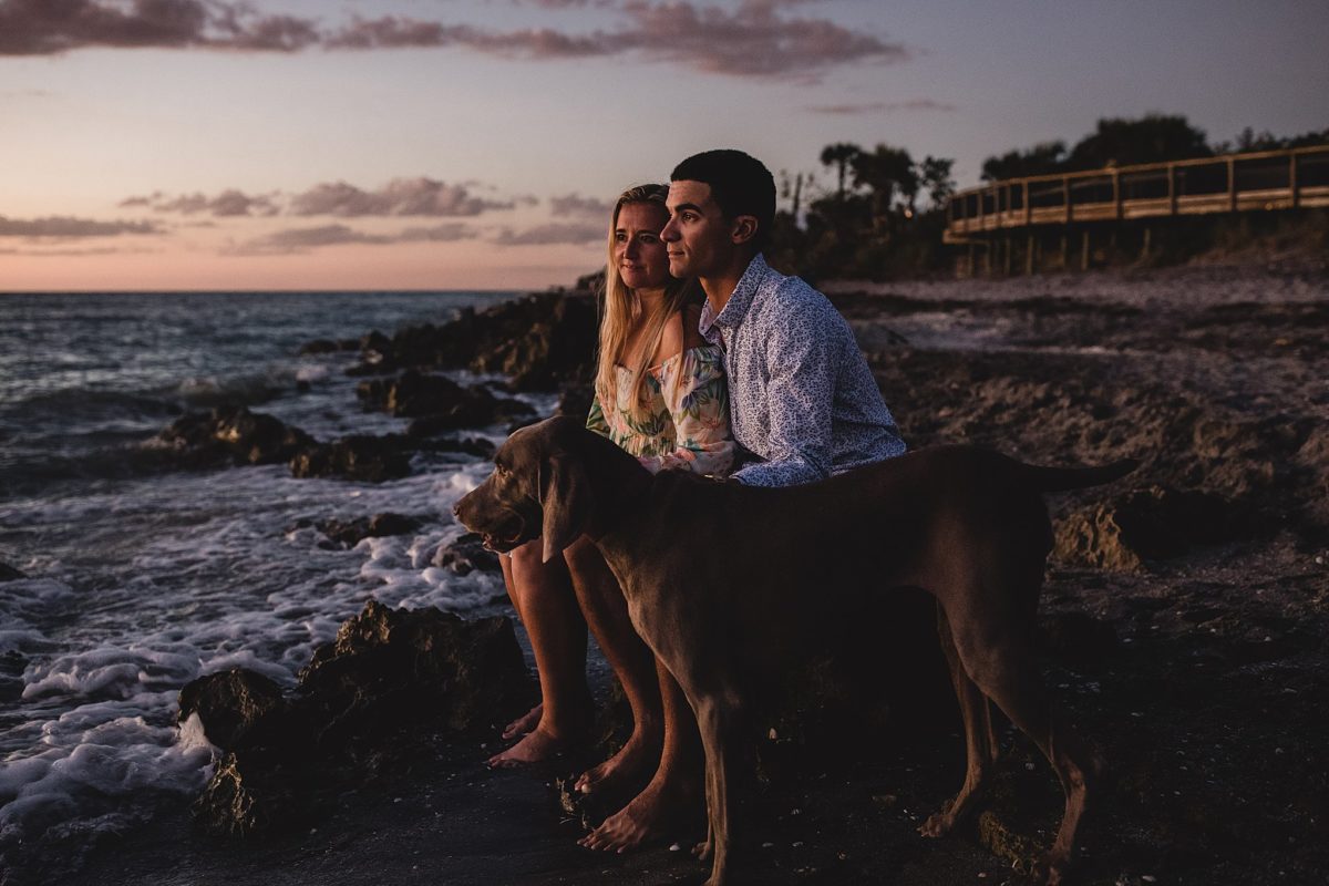 couple sitting with their dog at caspersen beach looking at the sunset, engagement photos by juliana montane photography