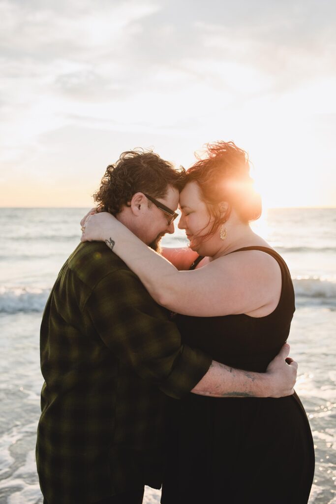 Couple with their foreheads together on the beach with the sun setting behind them after getting engaged. Photographed by Juliana Montane Photography.