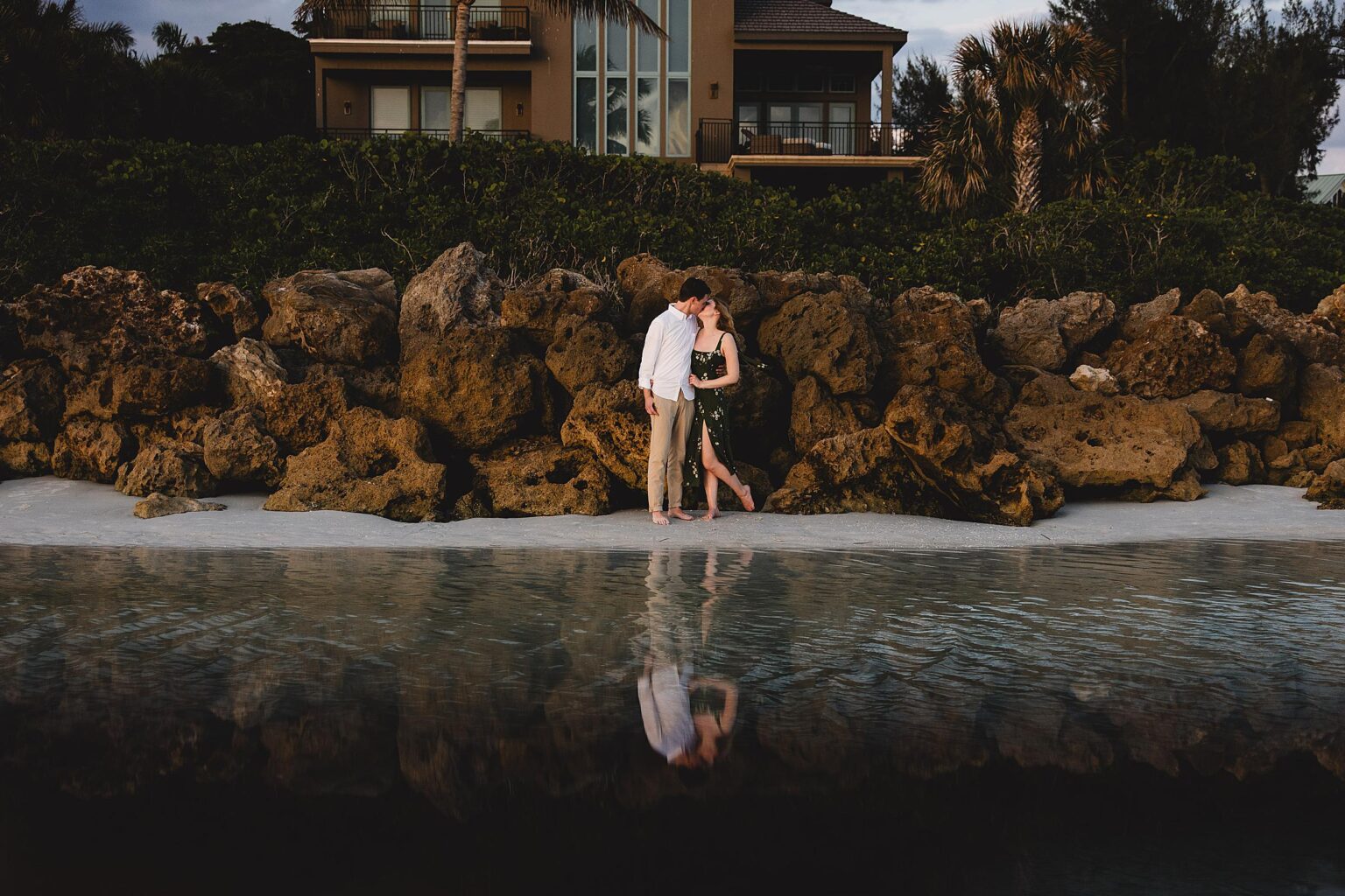 Couple kissing in front of rocks along the beach during an engagement session on Siesta Key Beach. Photographed by Juliana Montane Photography.