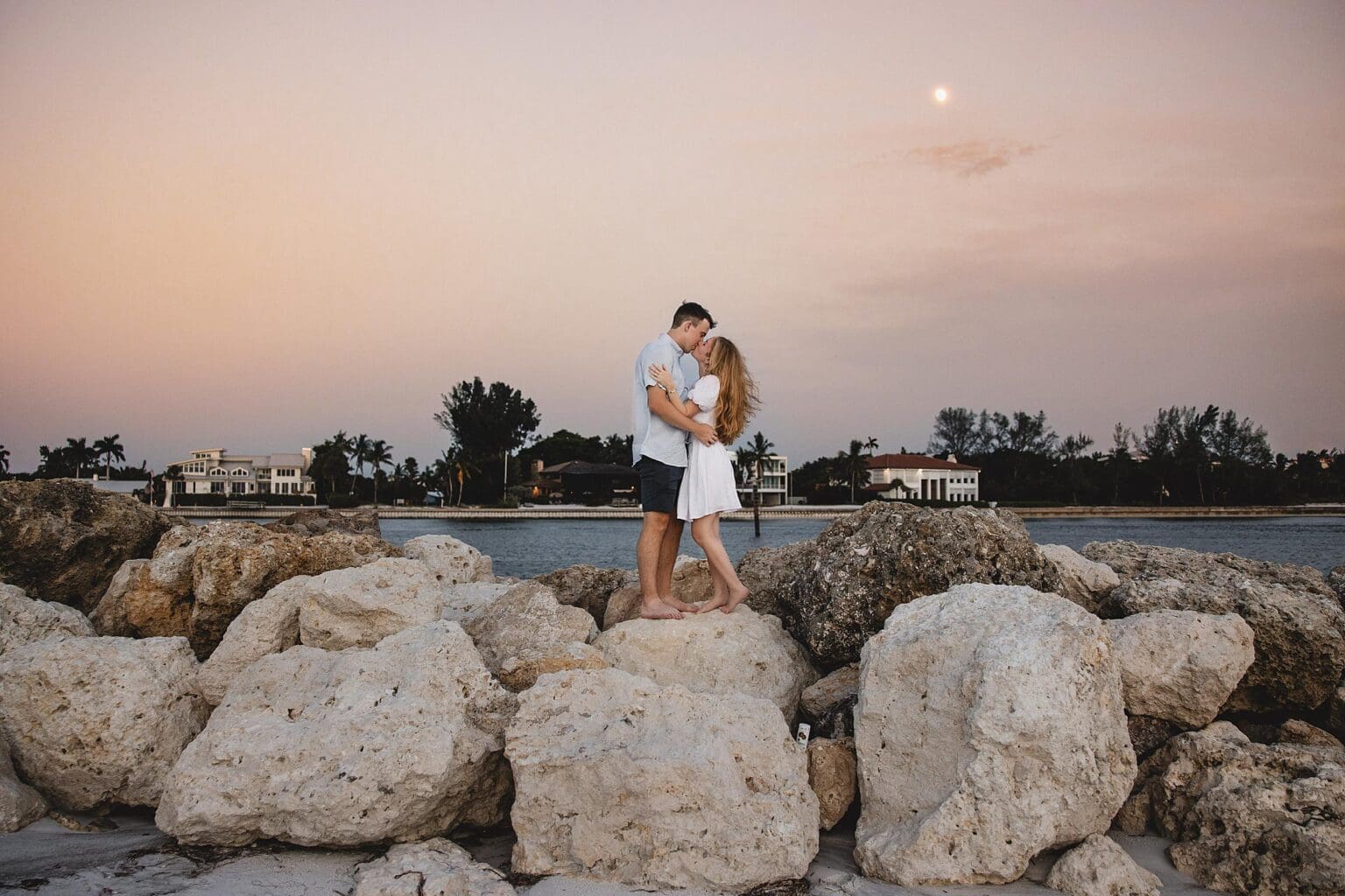 Newly engaged couple kissing on rocks on Longboat Key Beach after their sunset surprise proposal in Sarasota, FL photographed by Juliana Montane Photography